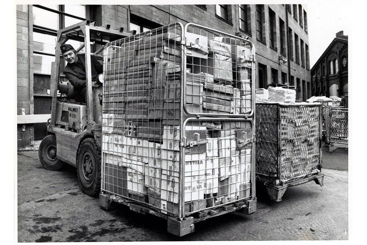 Postin 1970-luvun työasussa oleva mies siirtää paketteja täynnä olevaa tavarakärryä haarukkatrukilla.