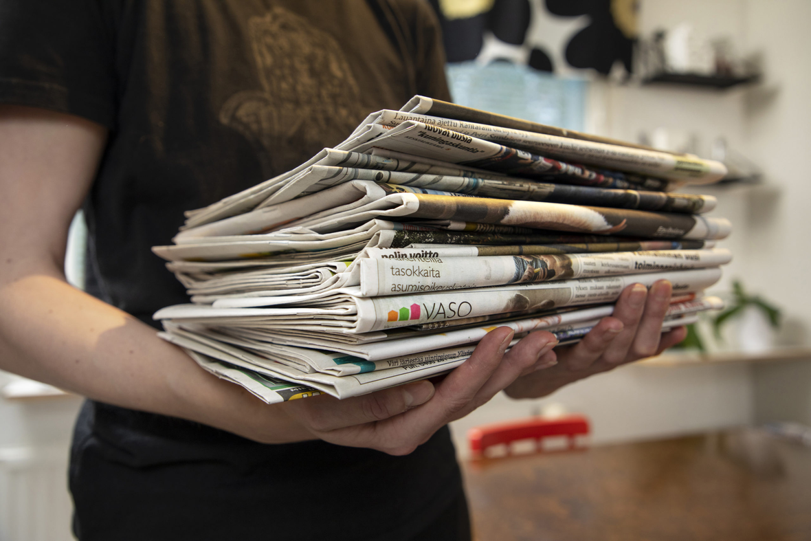 Ihminen kantaa keittiössä sylissään sanomalehtipinoa.