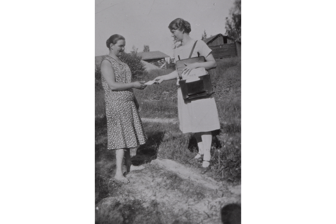 Mustavalkoisessa kuvassa nainen oikealla vaaleassa mekossa ja nahkalaukku olalla ojentaa kirjettä toiselle naiselle.