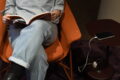Henkilö istuu nojatuolissa kirjaa lukien, vieressään puhelin pöydällä latautumassa.